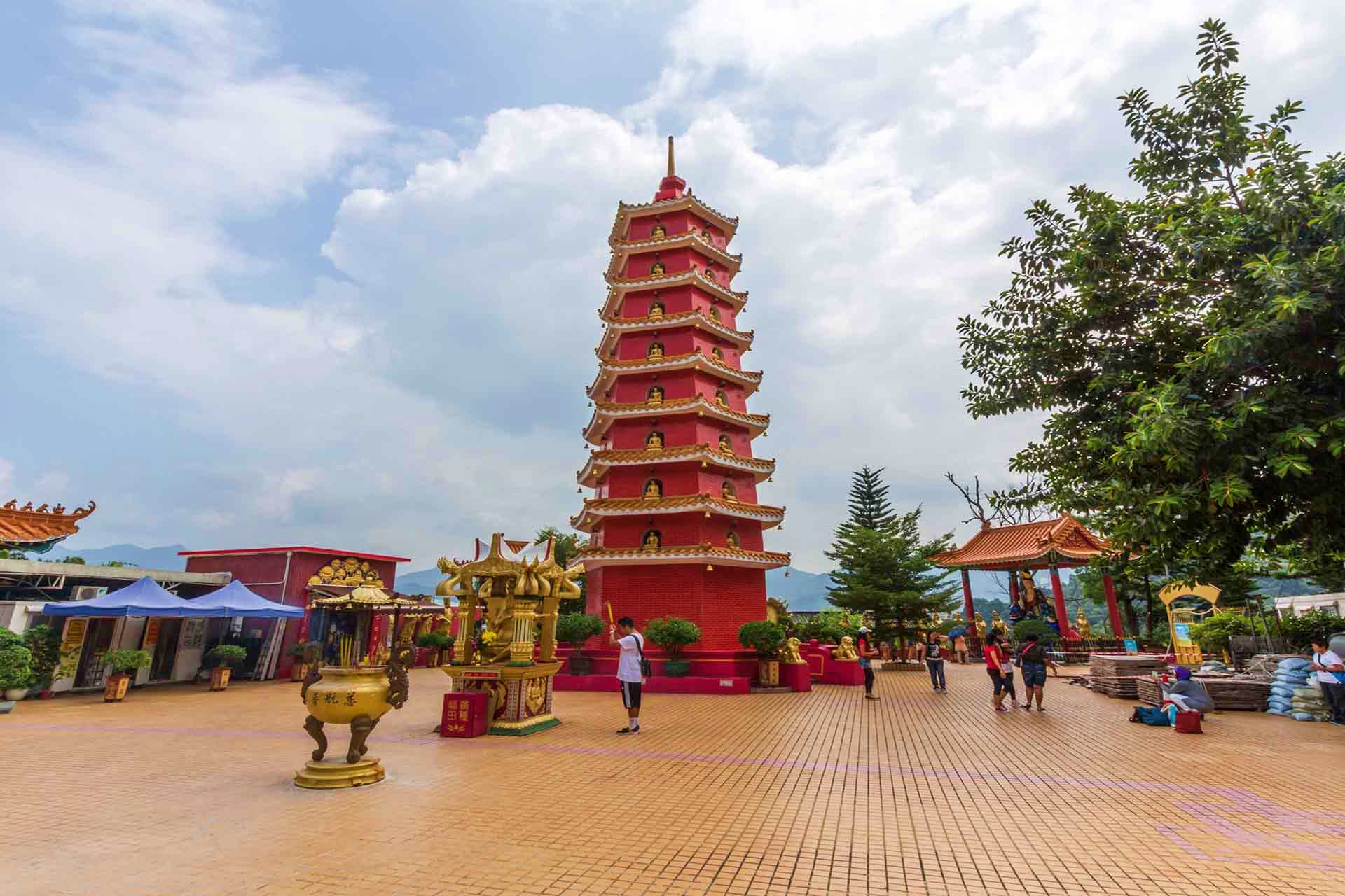 Red and gold Chinese pagoda tower at temple courtyard with visitors and ornamental urns under cloudy sky